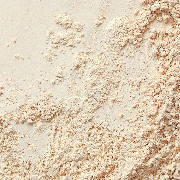 Close-up of loose, beige botanical powder scattered unevenly across a surface. The texture appears fine with some clumps spread throughout, creating a natural and organic pattern.