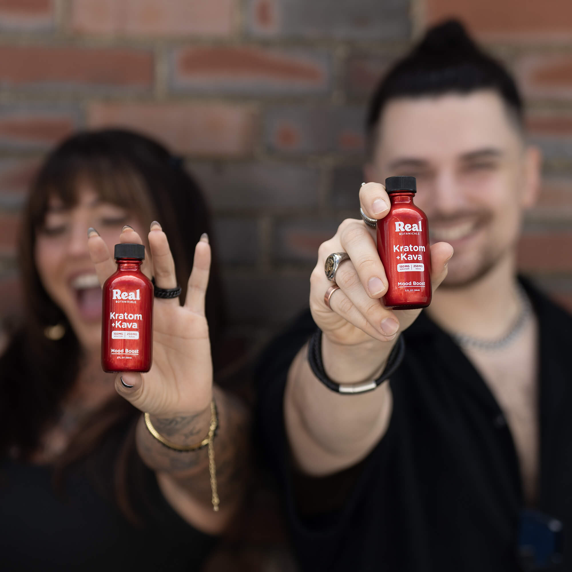 Two people holding red bottles labeled 'Real Kratom Kava' against a brick wall.