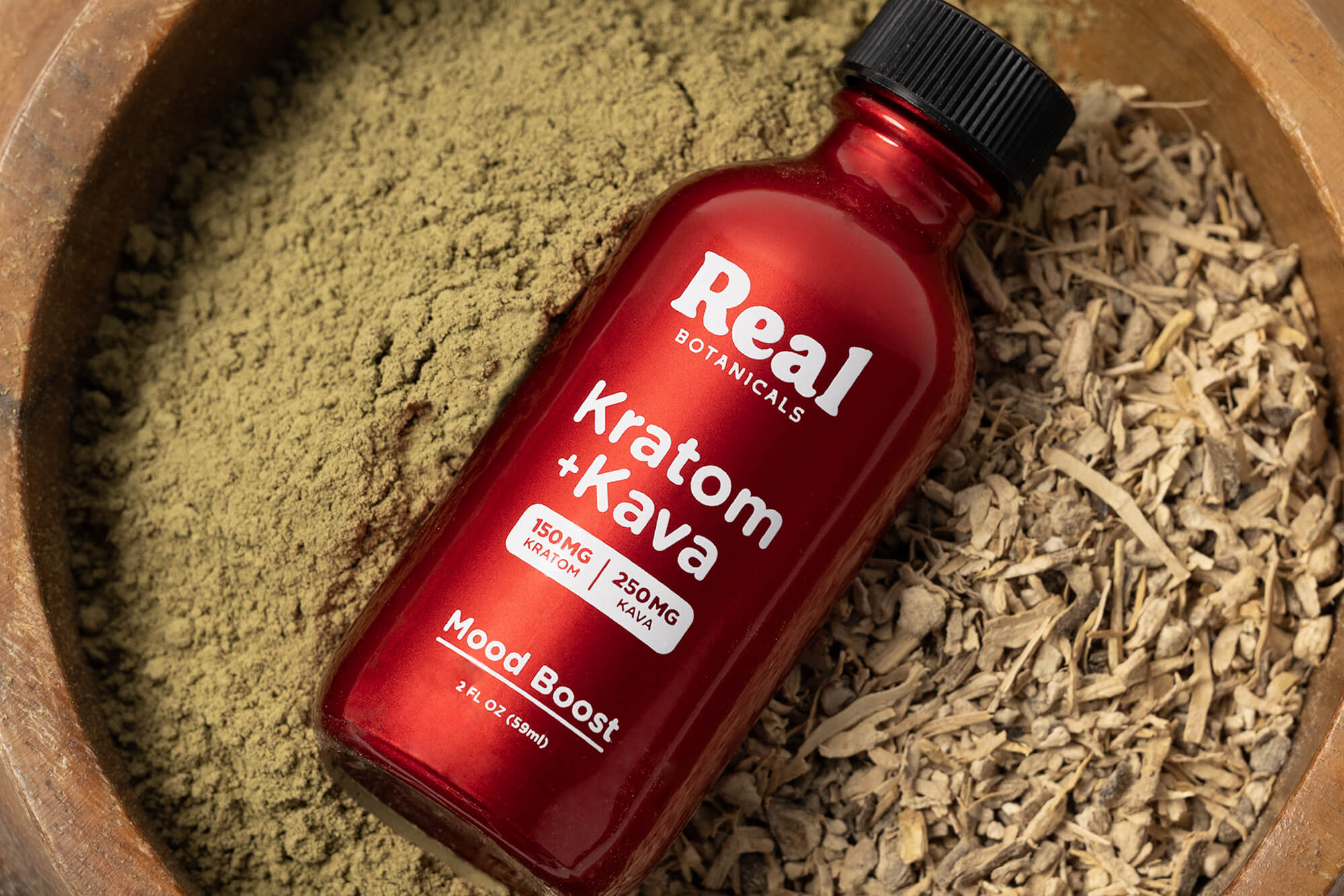 Red bottle labeled 'Real Botanicals Kratom + Kava' on a wooden surface with dried herbs.