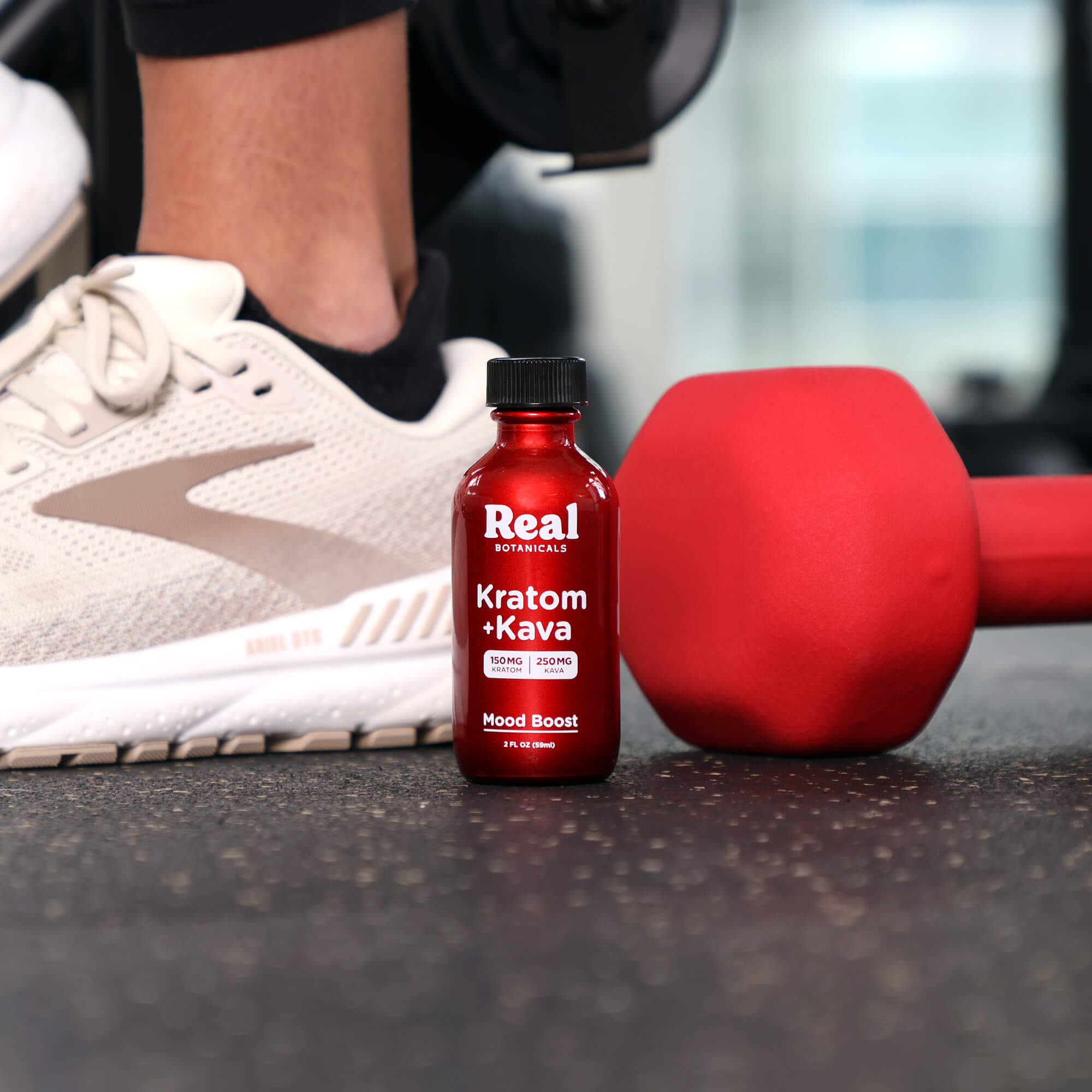 Red bottle of Real Supplements Kratom Kava next to a red dumbbell and a shoe on a gym floor.