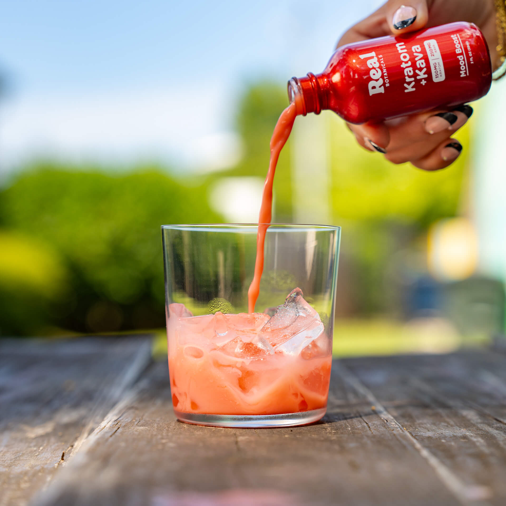 Red bottle being poured into a glass of pink liquid with ice on a wooden table outdoors.