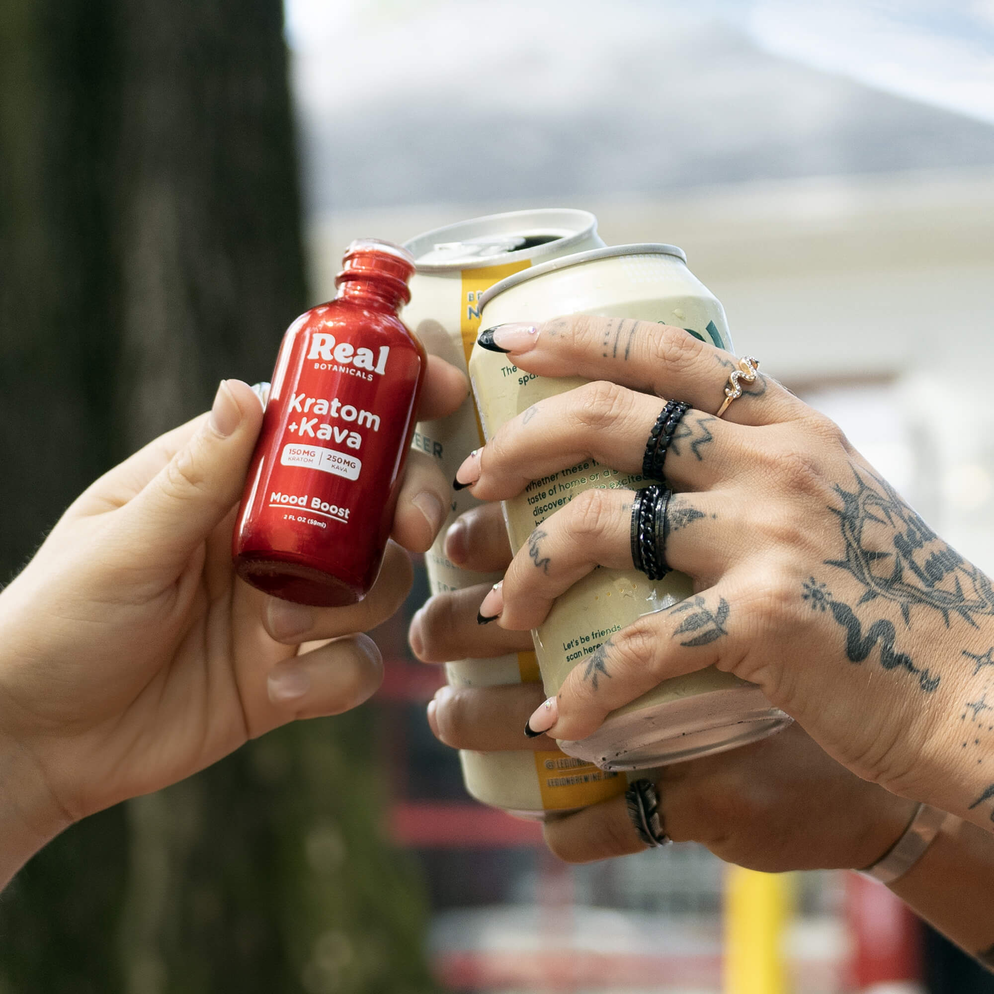Two hands holding a red bottle labeled 'Real Kratom Kava' and a can, with tattoos visible on the hands.