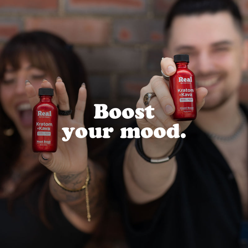 Two people smiling and holding small red bottles labeled Real Botanicals Kratom + Kava with a brick wall backdrop. The text Boost your mood is centered in bold white letters.